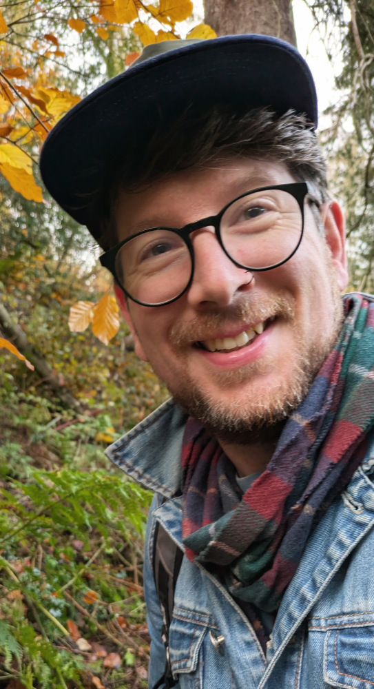 Smiling man with glasses and a cap among autumn foliage.